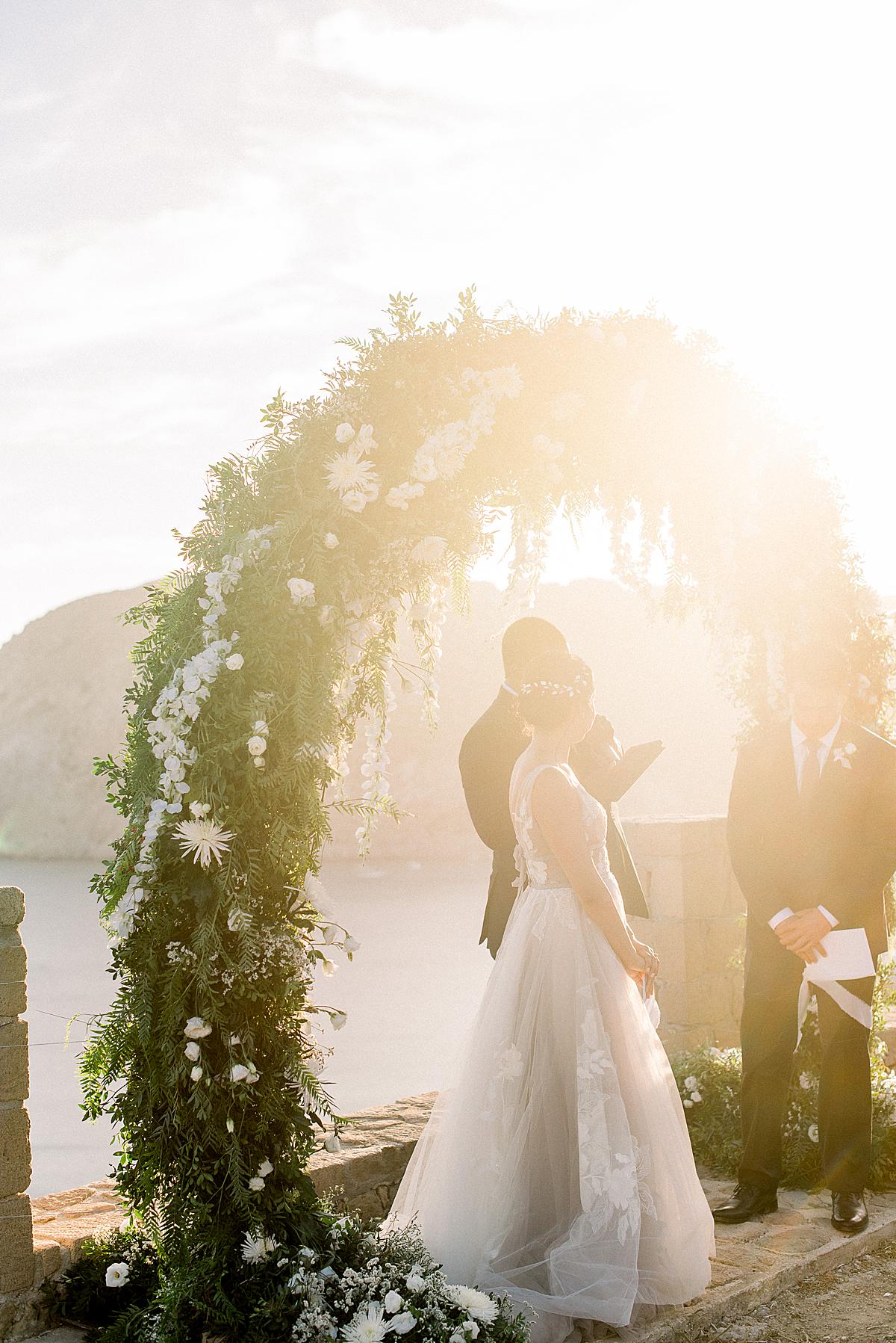 bride and groom under their sunkissed arch of flowers at their milos wedding