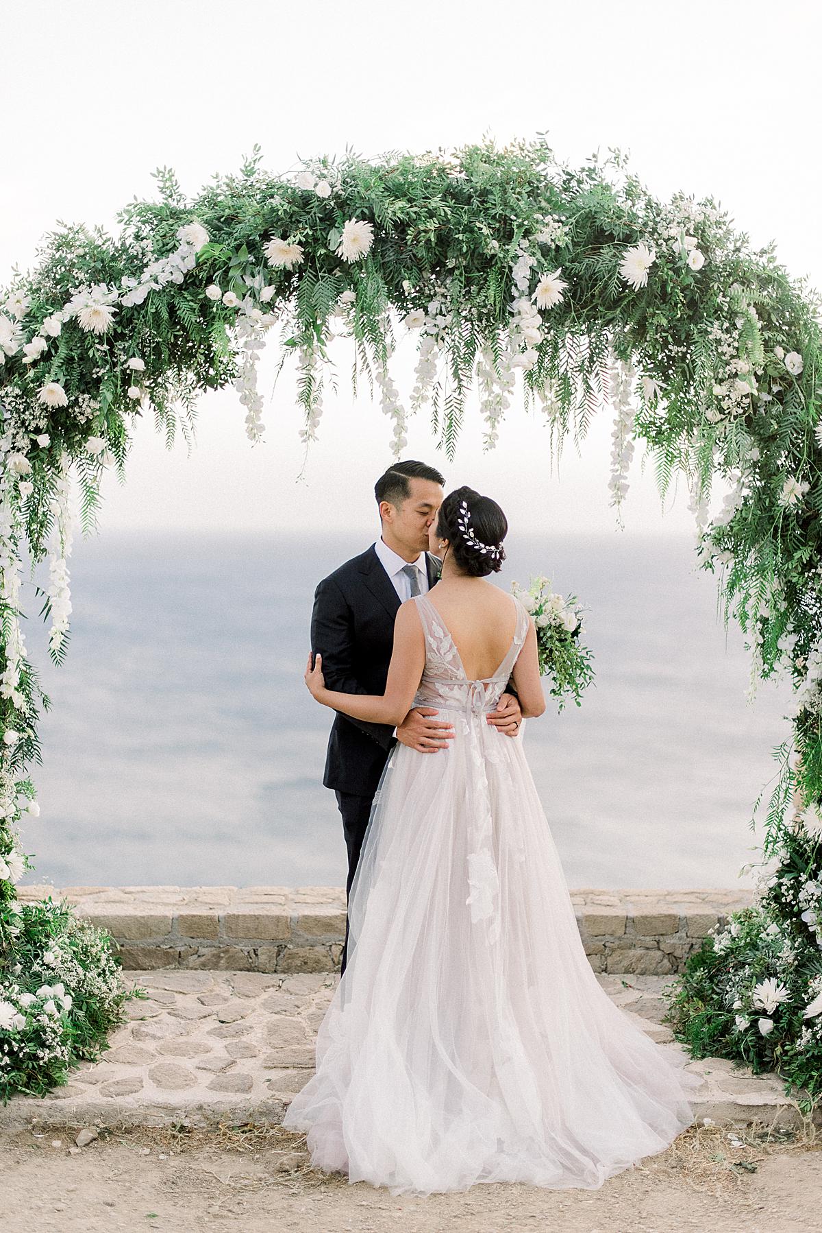 bride and groom under their sunkissed arch of flowers at their milos wedding