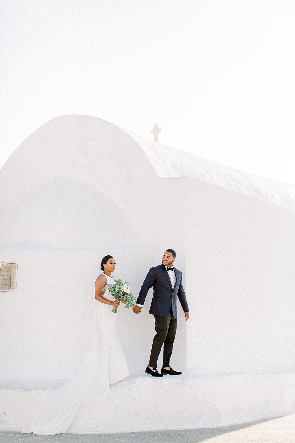 groom in hand with bride in front of washed out white church in santorini